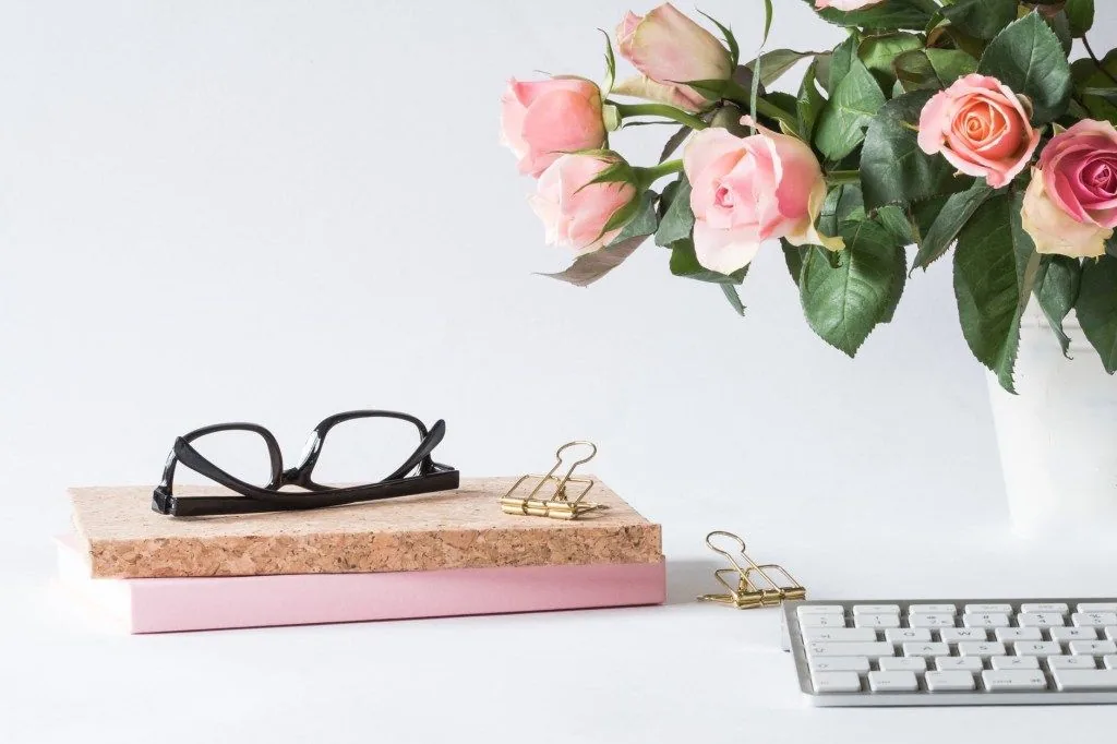 Glasses, books, keyboard, and pink roses on a white desk