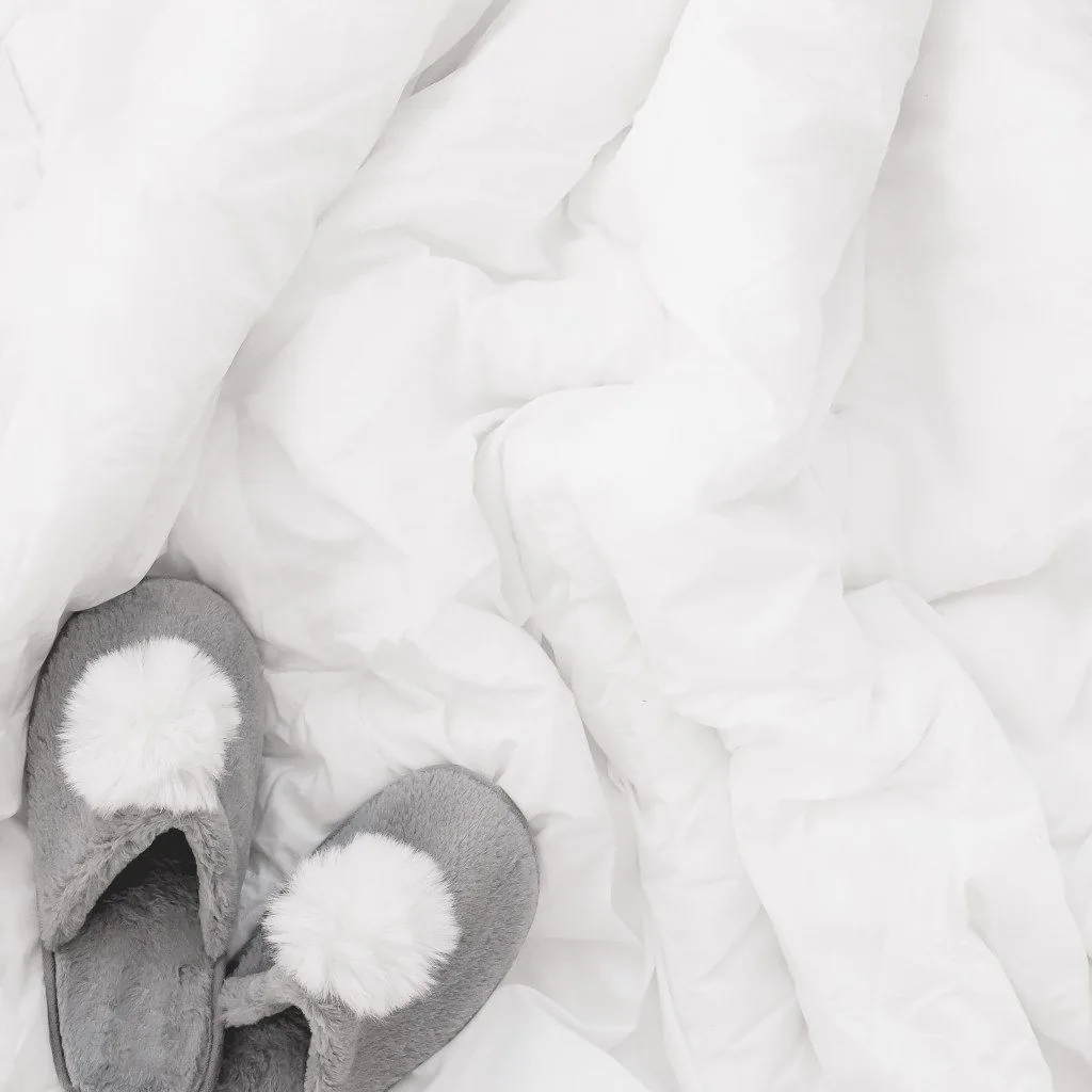 Gray slippers with white pom-poms on rumpled white bedding